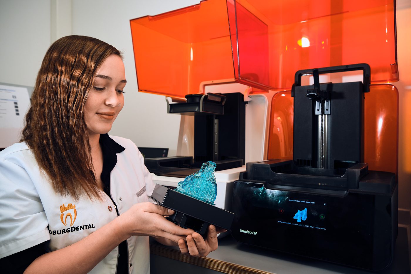 Mitarbeiterin im Dentallabor hält ein 3D-gedrucktes Bauteil vor einem 3D-Drucker