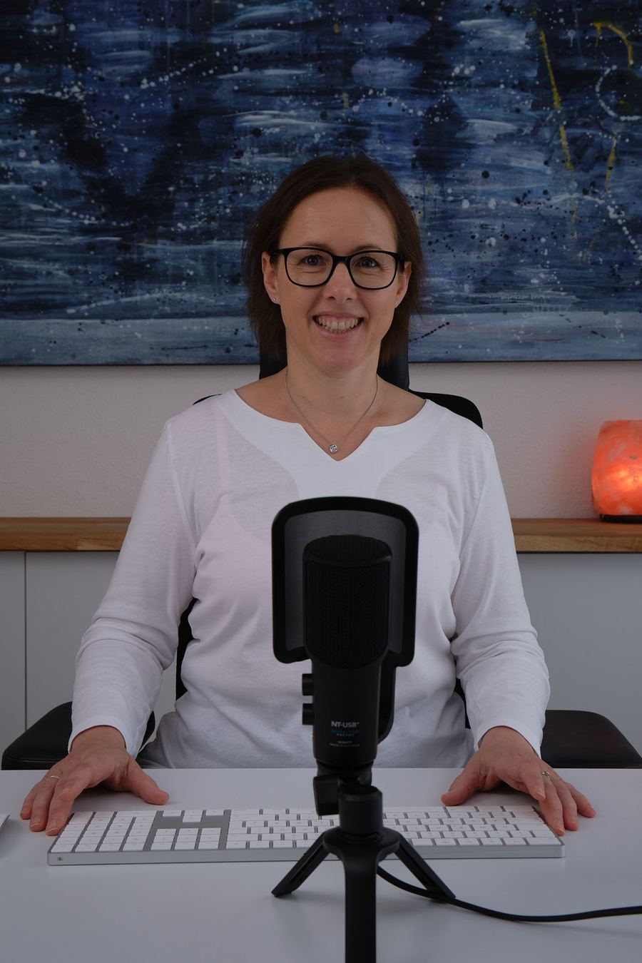 Frau mit Brille sitzt vor einem Mikrofon und einer Tastatur am Schreibtisch