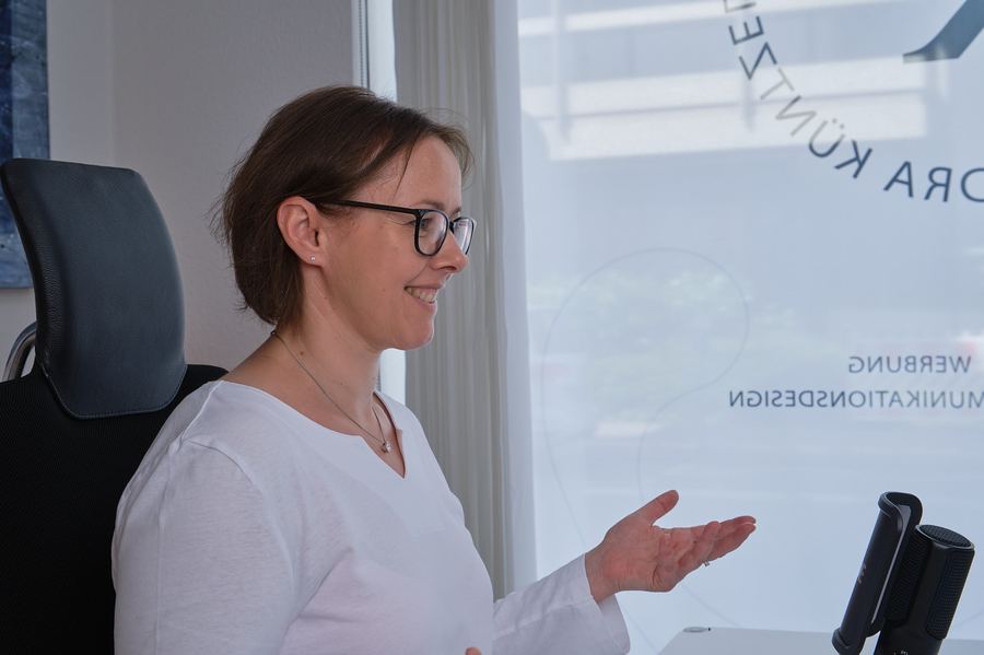 Frau mit Brille im Gespräch, gestikuliert lächelnd an einem Bürofenster