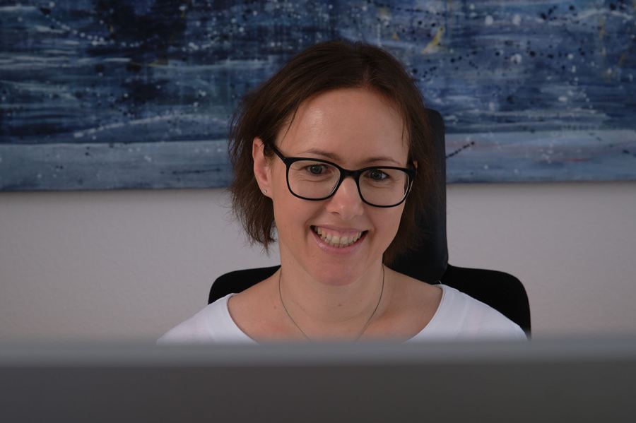Frau mit Brille lächelt an ihrem Arbeitsplatz in die Kamera